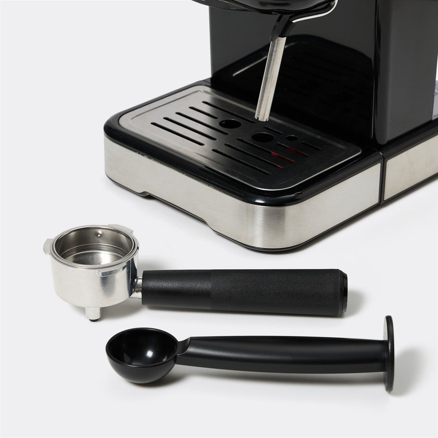 A close-up of a coffee machine with its accessories laid out in front, including a stainless steel portafilter, a black scoop, and a black cleaning brush, on a light gray background.