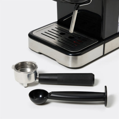 A close-up of a coffee machine with its accessories laid out in front, including a stainless steel portafilter, a black scoop, and a black cleaning brush, on a light gray background.