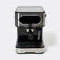 A compact espresso machine with a brushed stainless steel body and black accents, featuring a portafilter and steam wand, photographed on a light gray background.