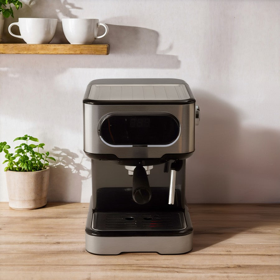 A compact espresso machine with a brushed silver and black finish, set on a wooden countertop against a light wall. A small potted green plant and a wooden shelf with white mugs are visible in the background.