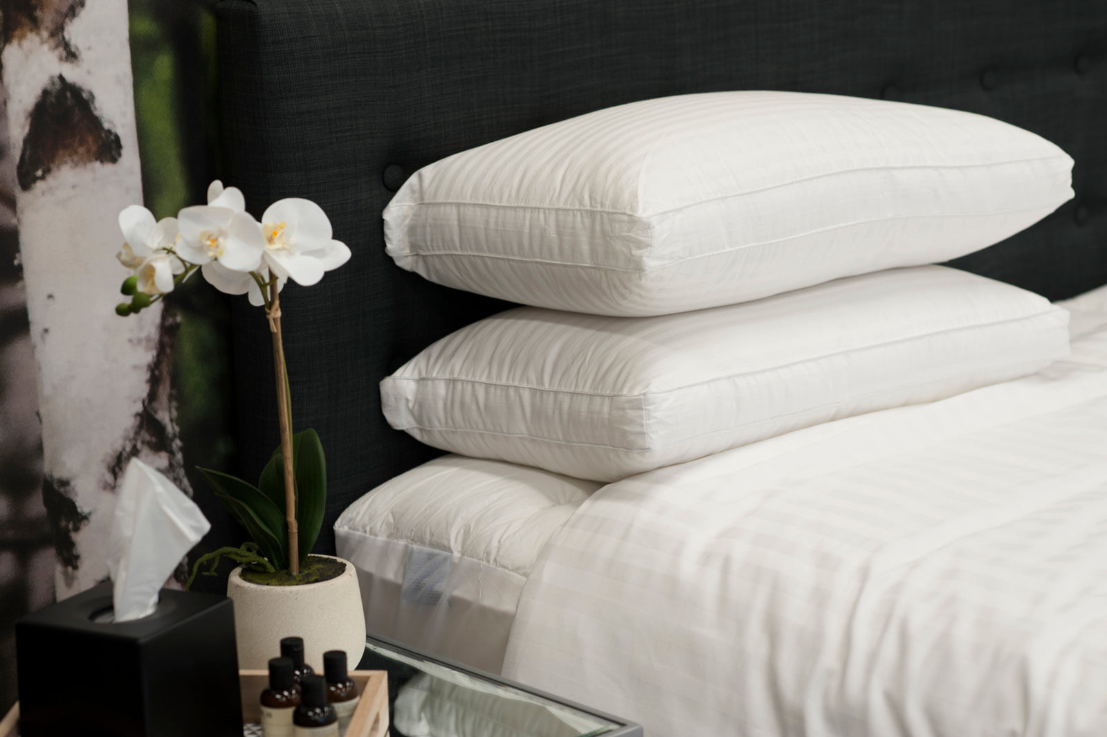 Stack of white pillows on a bed with a flower arrangement and small table in the foreground.