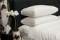 Stack of white pillows on a bed with a flower arrangement and small table in the foreground.