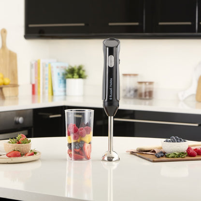 Black immersion blender on a kitchen counter with fruits and a glass.