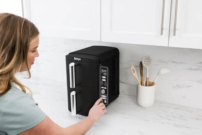 Woman using a Ninja foodi appliance on a kitchen counter with utensils nearby.