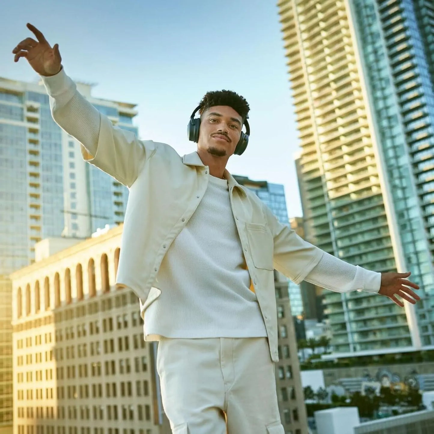 Man in a beige jacket and pants standing in an urban setting with tall buildings.