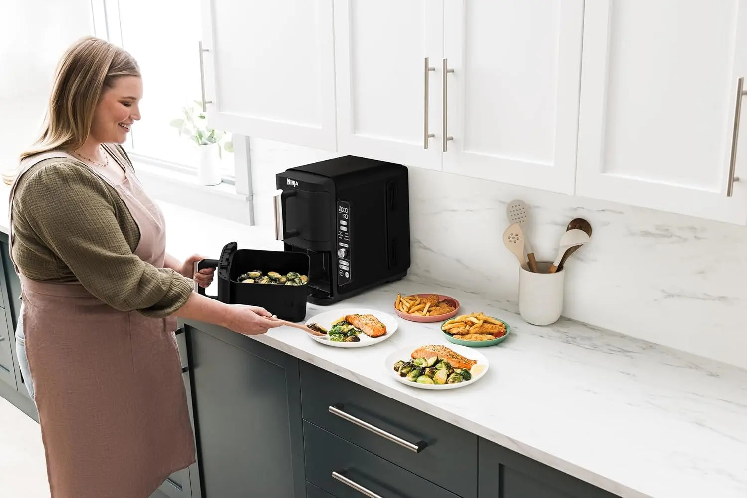 Woman in a kitchen using a countertop appliance with plates of food nearby.