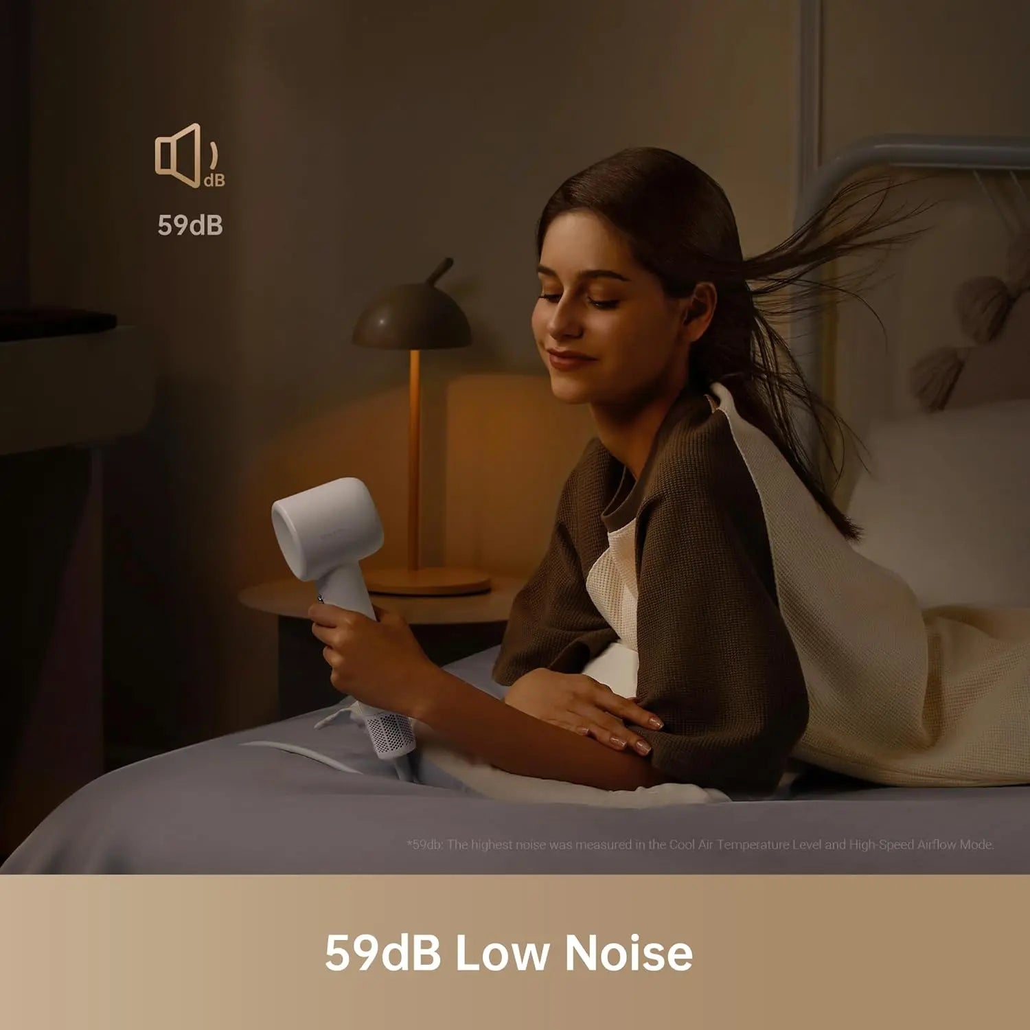 Woman using a hair dryer with 59dB low noise indicator in a bedroom setting.