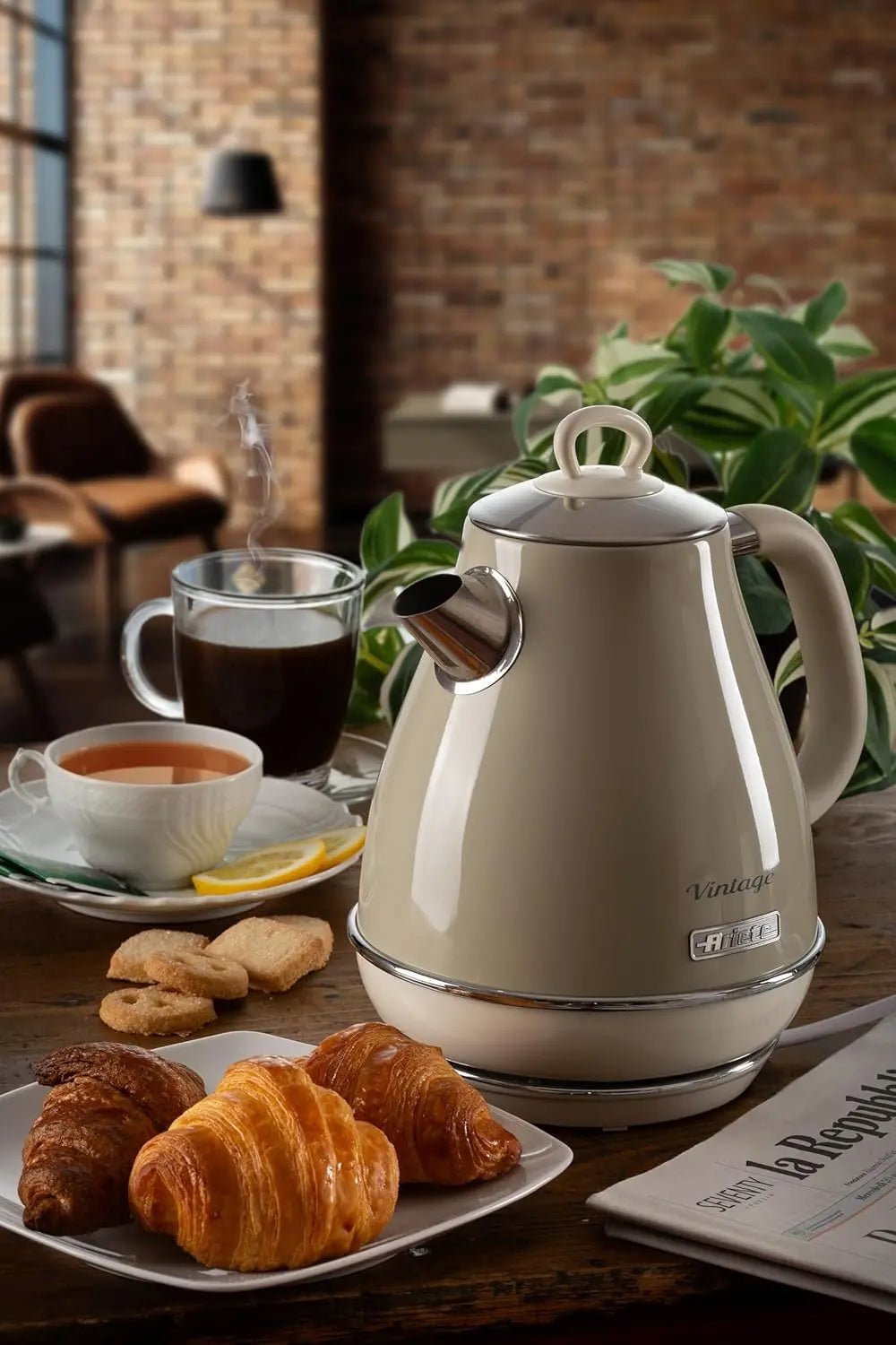 White electric kettle on a table with croissants, coffee, and a newspaper in a cozy setting.