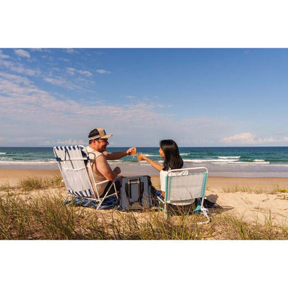 A man and a woman sit on beach chairs on a sandy dune at the ocean, clinking drinks. A gray portable cooler sits between them, with blue and white striped beach chairs and a light blue lounge chair nearby.