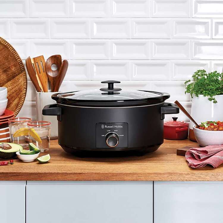 Black slow cooker on a kitchen counter with utensils and ingredients.