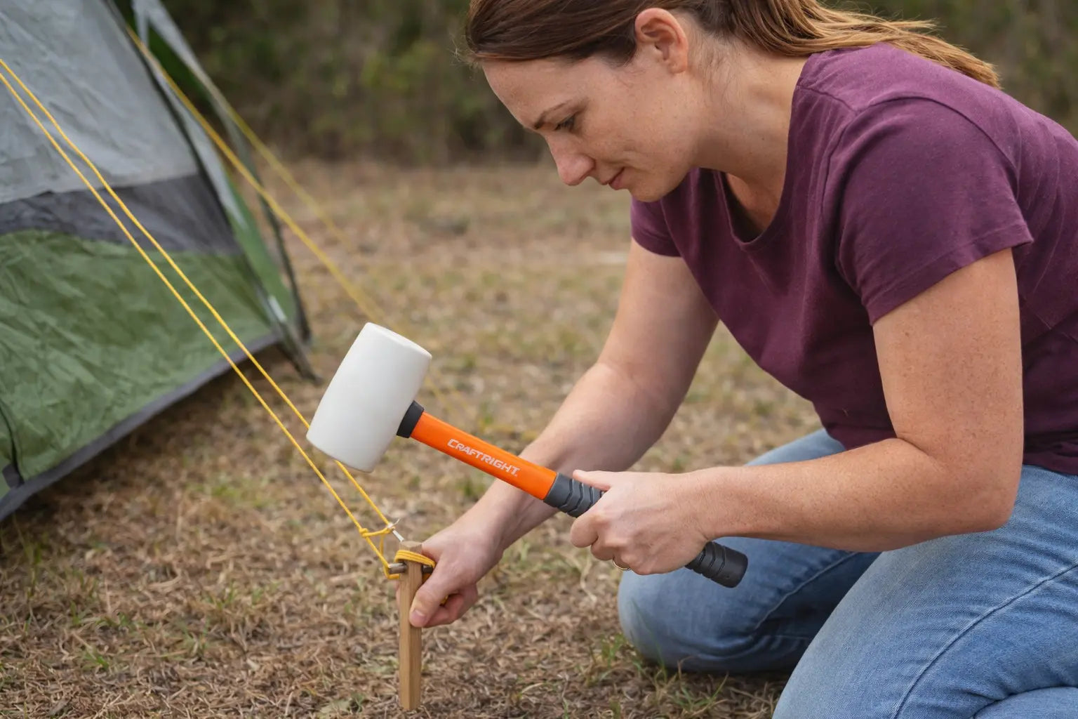 Woman setting up a camping hammock with a hammer near a tent.