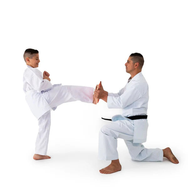 A man and a young boy in white martial arts uniforms practice a kick in a studio setting against a plain white background.