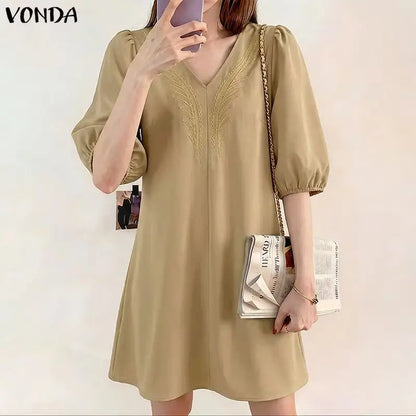 Woman wearing a beige dress holding a phone and a clutch against a white background