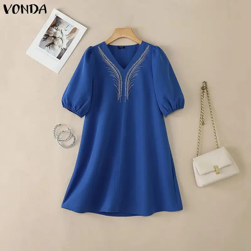 Blue dress with embroidery on a beige background, accompanied by a white handbag and photo frame.