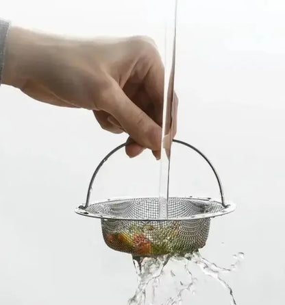 Hand holding a metal mesh strainer over a glass of water with fruits inside, on a white background.