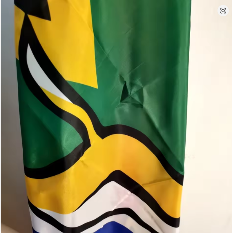 A close-up of a flag with a green upper field, a yellow chevron, black border lines, white and black bands, and a blue section near the bottom, photographed indoors against a light wall.