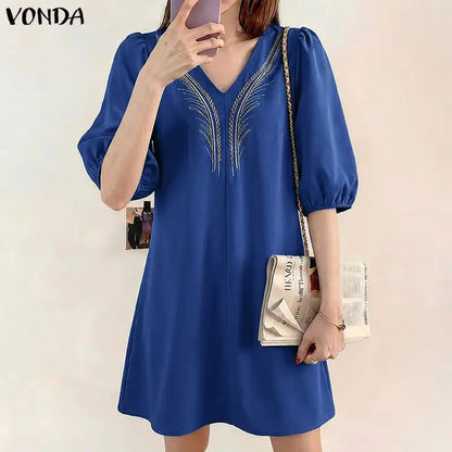 Woman wearing a blue dress with embroidery, holding a clutch, against a plain background.