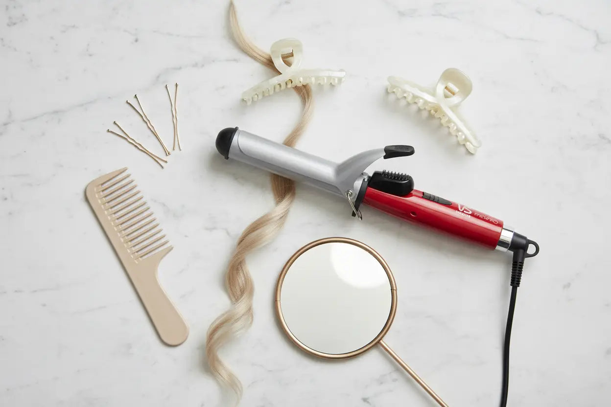 Hair styling tools including a comb, hair straightener, and hair curler on a marble surface.