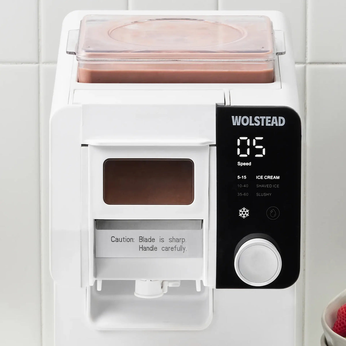 A white Wolstead ice cream maker with a black digital control panel and a clear container filled with pink ice cream, set against a white tiled background.