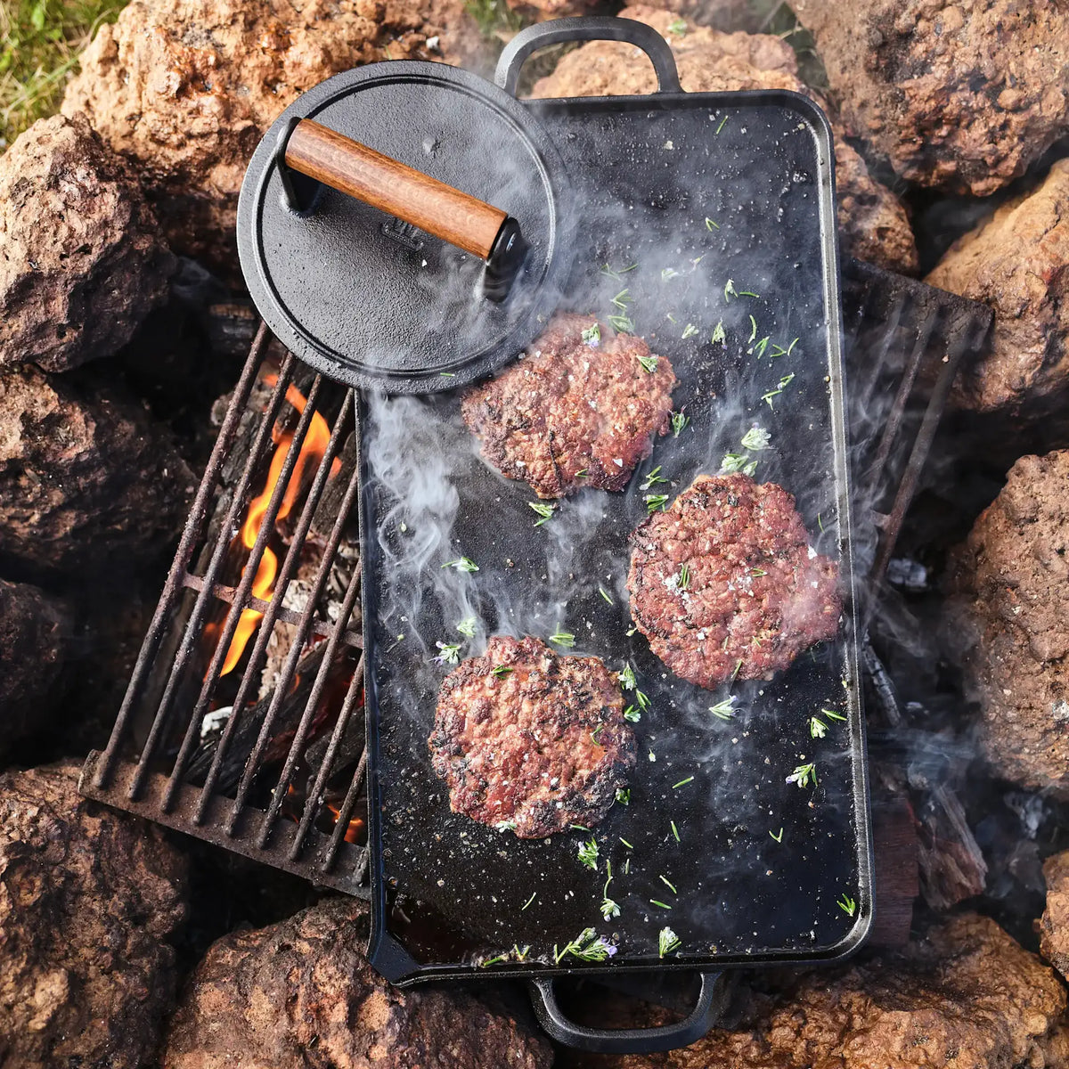 A black cast iron outdoor griddle with a lid, shown cooking burger patties over an open campfire surrounded by rocks. The lid is placed on top of the griddle, and smoke rises from the cooking meat.