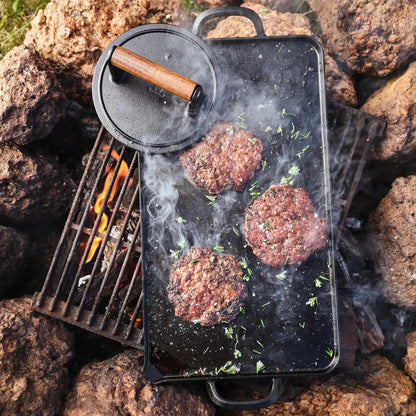 A black cast iron outdoor griddle with a lid, shown cooking burger patties over an open campfire surrounded by rocks. The lid is placed on top of the griddle, and smoke rises from the cooking meat.