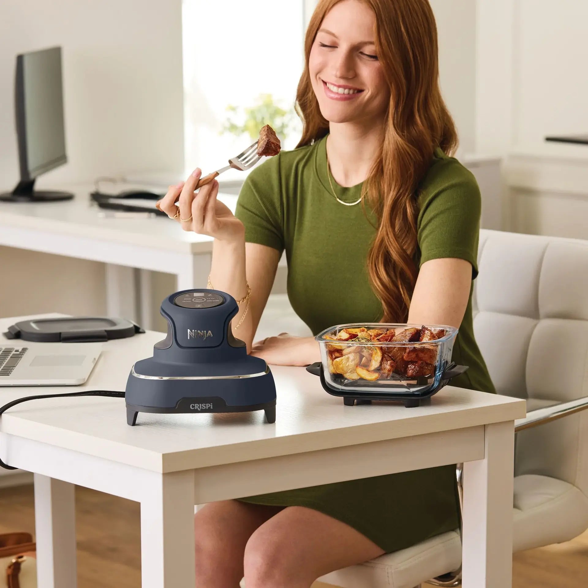 A woman sits at a white desk in a bright office setting, holding a fork with food while using a dark blue Ninja food processor and a clear glass baking dish on the desk.