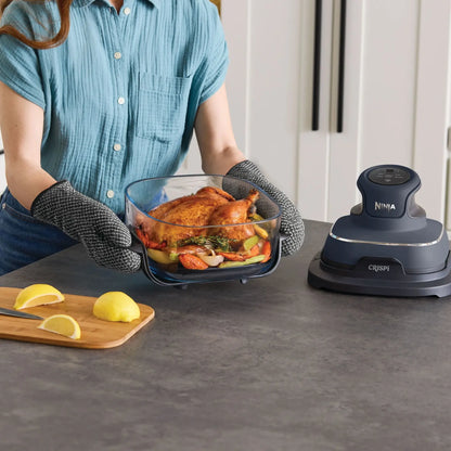 A person wearing a blue shirt and black oven mitts holds a glass roasting dish with a roasted chicken in a kitchen setting, next to a dark gray Ninja Crispi countertop oven.