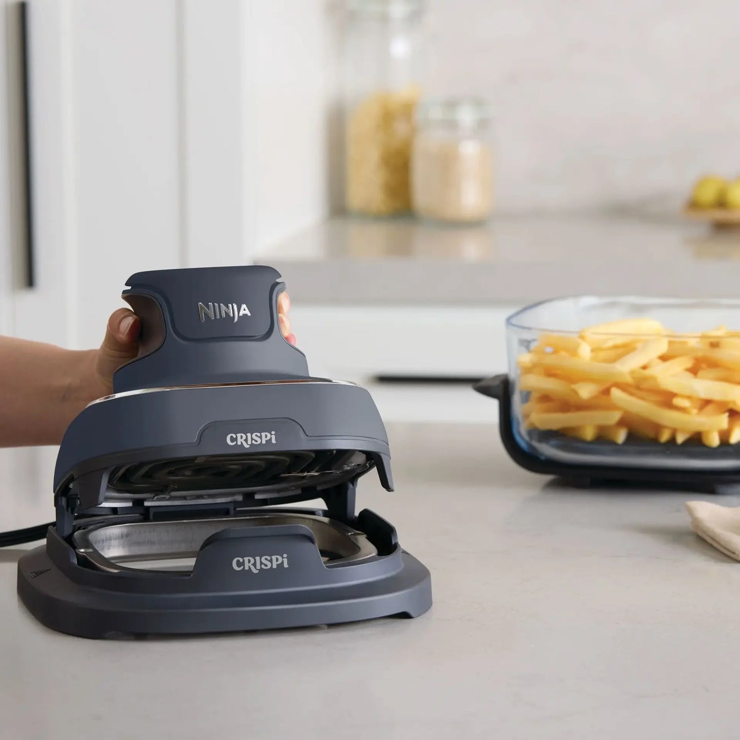 A hand is pressing down on a dark gray Ninja Crispi waffle maker on a light gray kitchen countertop, with a glass container of french fries blurred in the background.