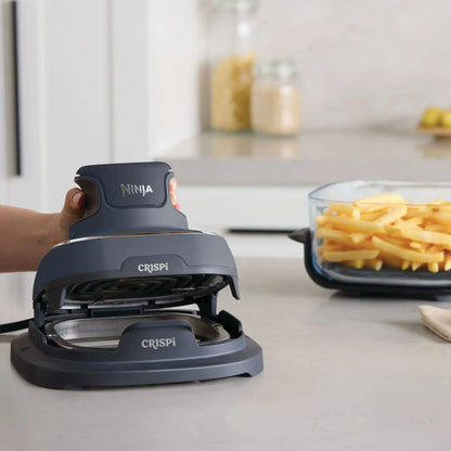 A hand is pressing down on a dark gray Ninja Crispi waffle maker on a light gray kitchen countertop, with a glass container of french fries blurred in the background.