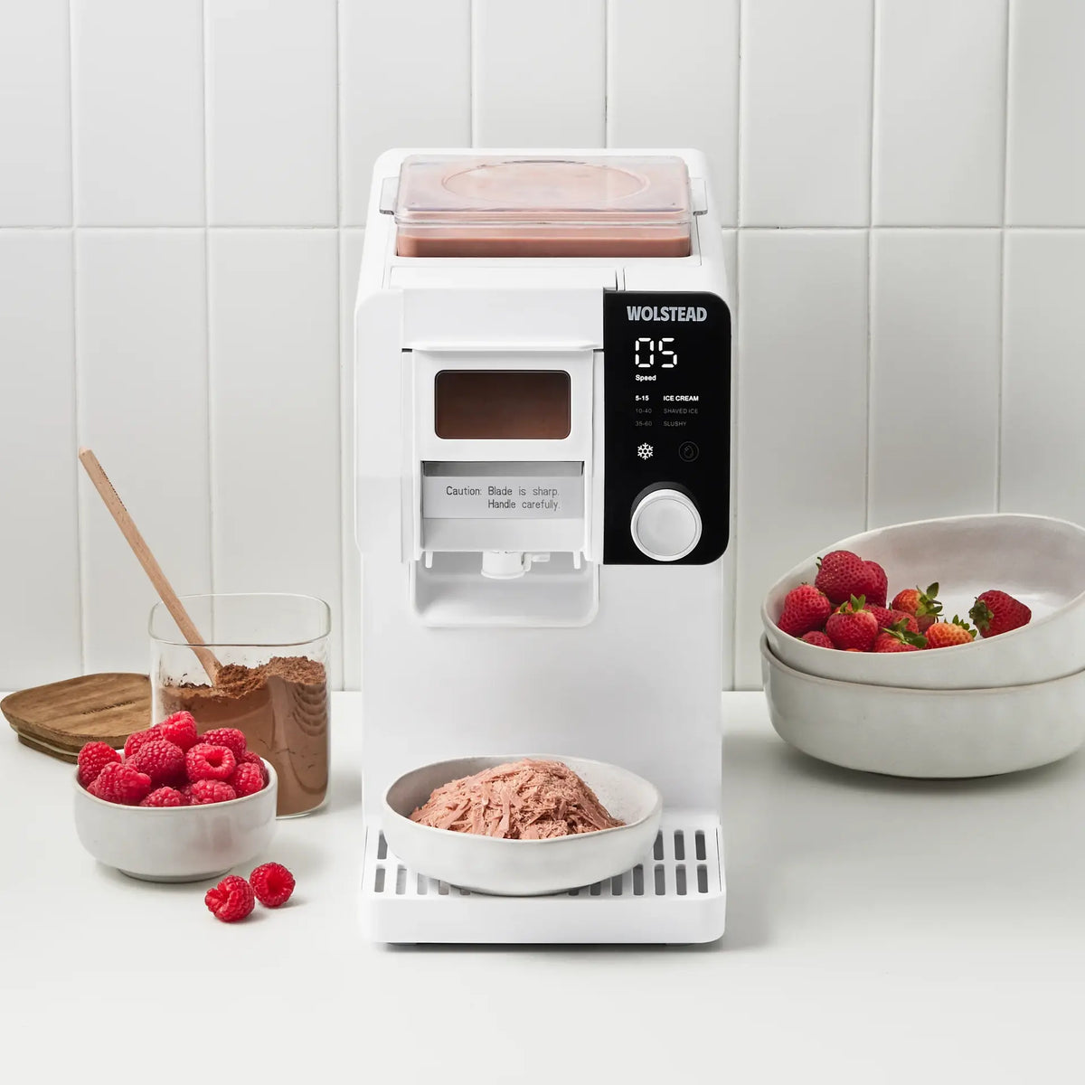 A white Wolstead ice cream maker sits on a white countertop in a kitchen setting, with bowls of raspberries, chocolate powder, and an ice cream cone in front of it. The background is a clean white tiled wall.