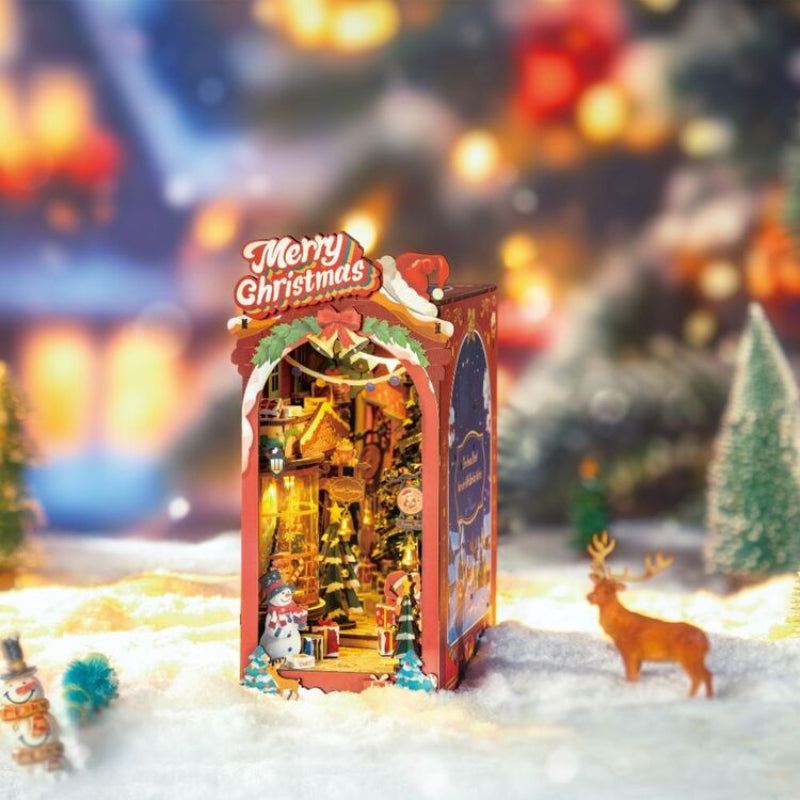 A Christmas-themed miniature display box with a glowing festive interior, topped with a sign reading 'Merry Christmas' and decorated with red and green holiday accents. The box is placed in a snowy scene with blurred holiday lights, trees, and small decorative figures around it.