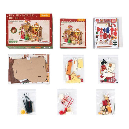 A DIY miniature house kit shown with its retail box, a smaller instruction card, a sticker sheet, and multiple clear bags containing wooden building pieces, decals, and accessories on a white background.
