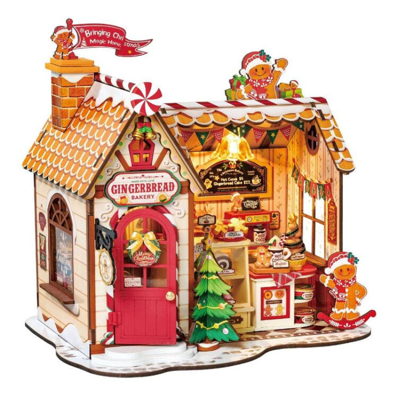 A detailed gingerbread-themed wooden toy house set with a bakery facade, decorated with gingerbread men figures, a small Christmas tree, candy cane accents, and warm glowing interior details, shown on a plain white background.